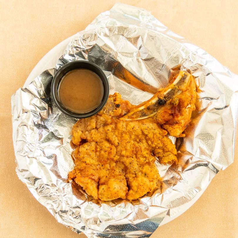Golden fried bone-in pork chop with a side of gravy, presented on aluminum foil.