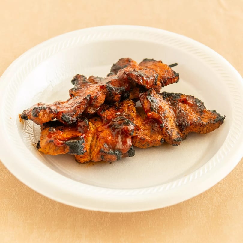 Charred and saucy grilled meat on a skewer, served on a white disposable plate.