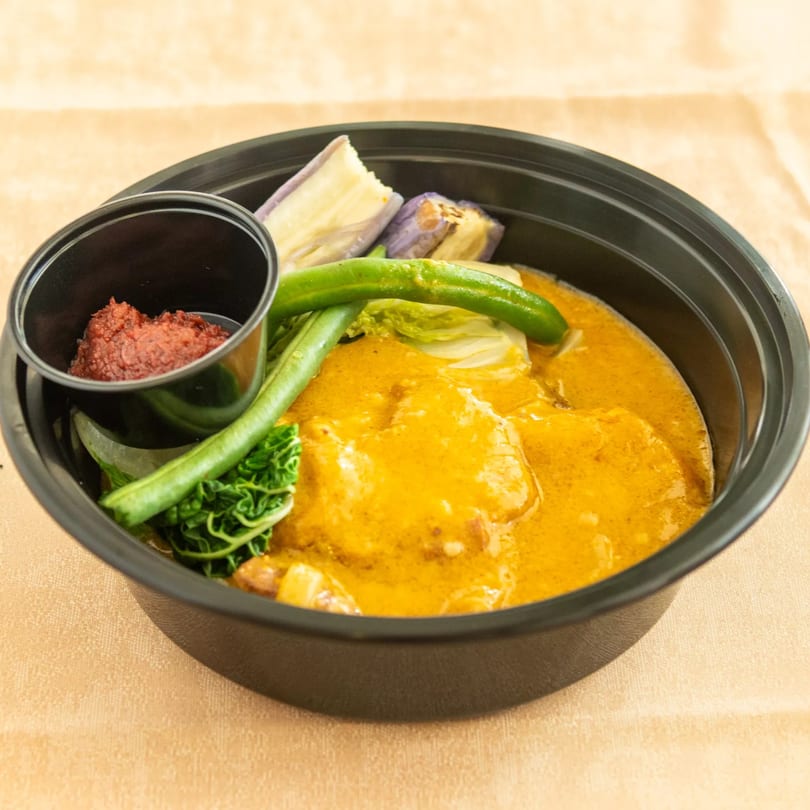 A black bowl of yellow curry with green beans, eggplant, cabbage, and a side of red paste.